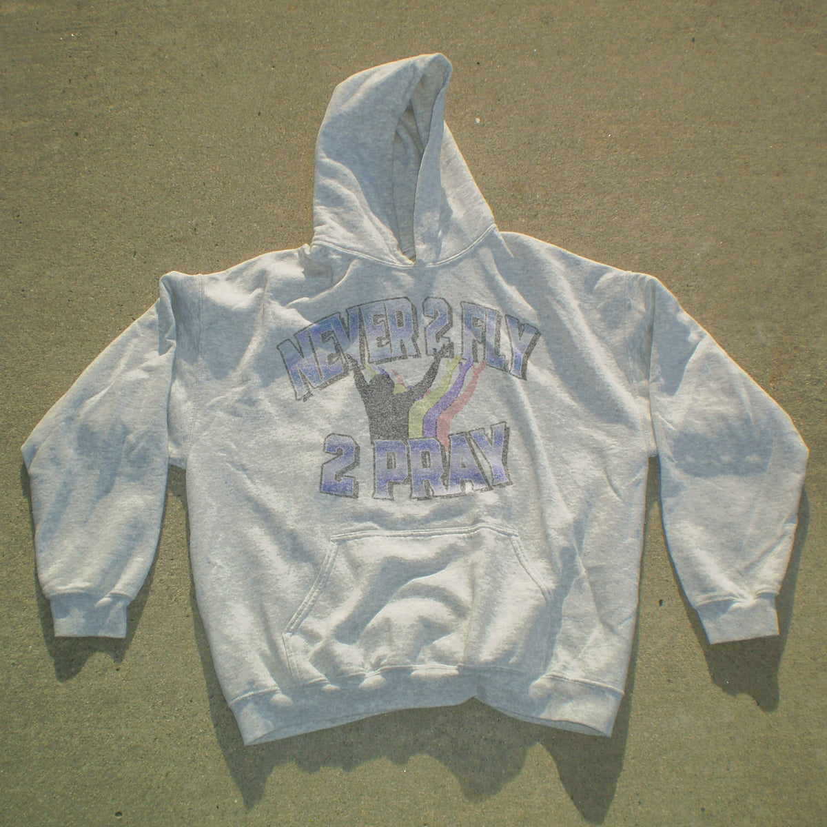 A light gray pullover hoodie laid flat on a concrete surface, featuring a distressed graphic on the chest that reads 'NEVER 2 HAY 2 PRAY' in purple and black with a purple ribbon design.