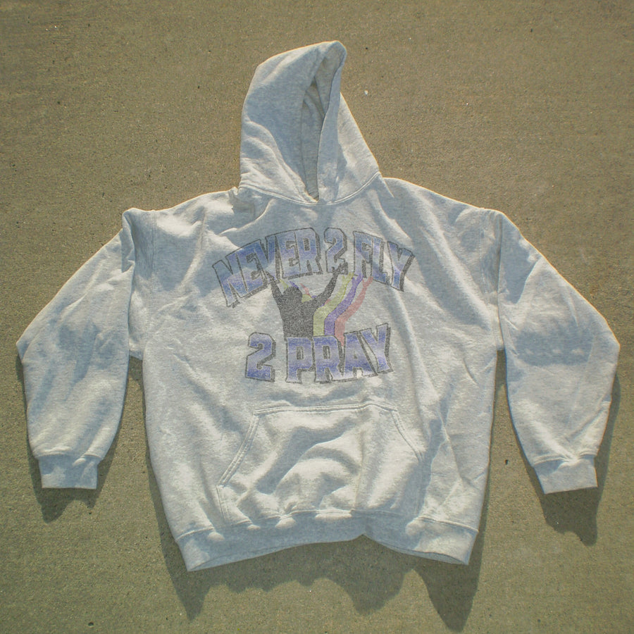 A light gray pullover hoodie laid flat on a concrete surface, featuring a distressed graphic on the chest that reads 'NEVER 2 HAY 2 PRAY' in purple and black with a purple ribbon design.
