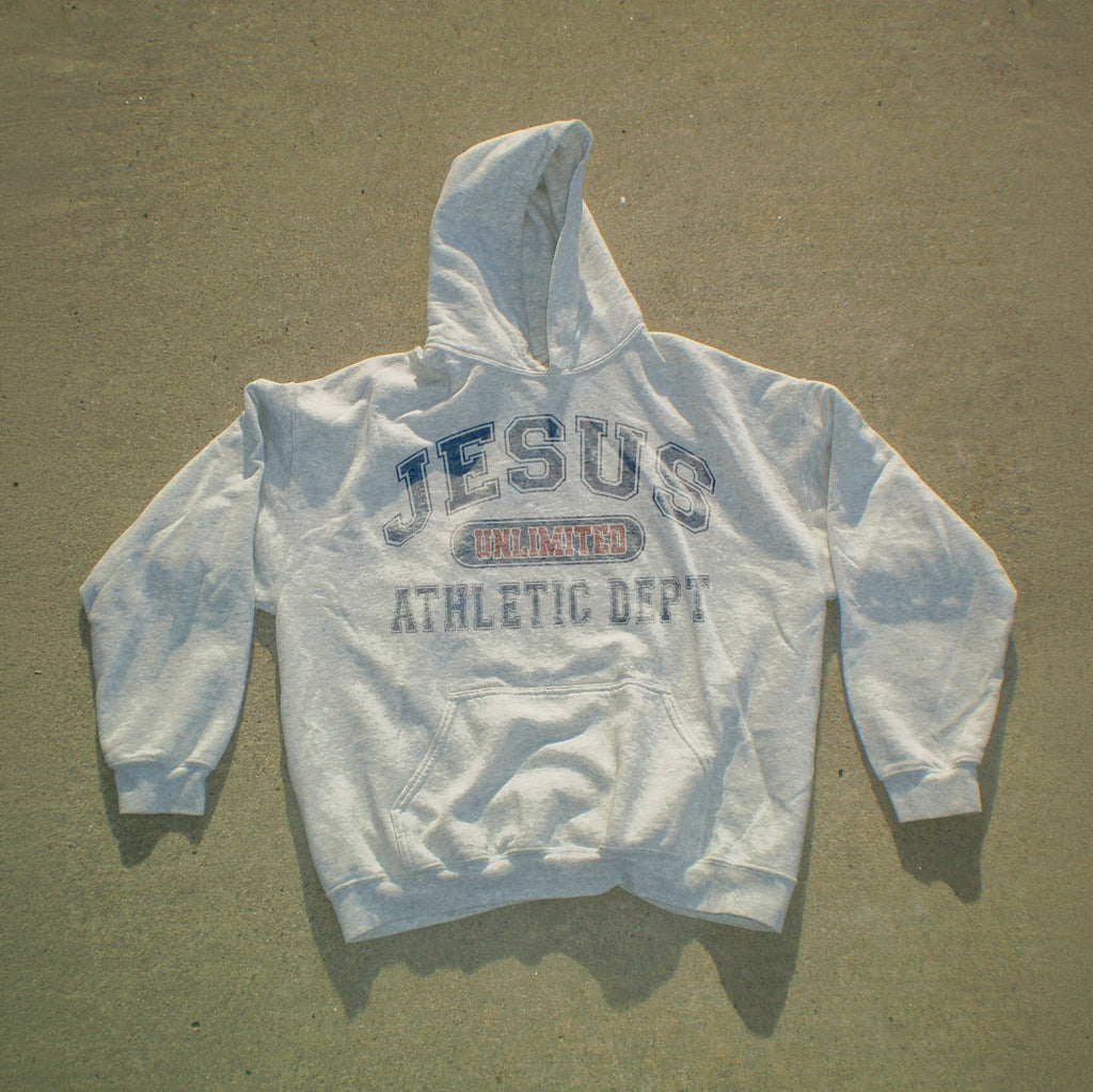 A light gray hooded sweatshirt laid flat on a textured concrete surface, featuring a distressed graphic on the chest that reads 'JESUS UNLAWED ATHLETIC DEPT' in blue and red.
