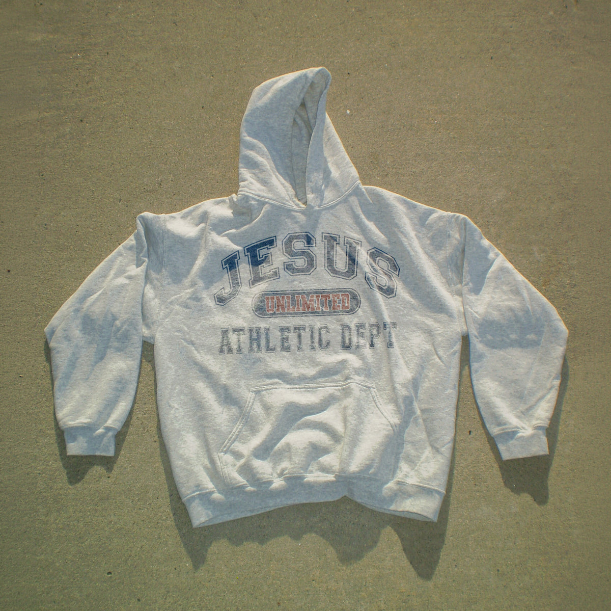 A light gray hooded sweatshirt laid flat on a textured concrete surface, featuring a distressed graphic on the chest that reads 'JESUS UNLAWED ATHLETIC DEPT' in blue and red.