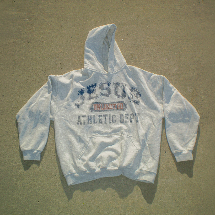 A light gray hooded sweatshirt laid flat on a textured concrete surface, featuring a distressed graphic on the chest that reads 'JESUS UNLAWED ATHLETIC DEPT' in blue and red.