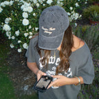 A woman outdoors wearing a dark gray baseball cap with gold embroidered text reading 'Breathe Easy Honey' and a gray graphic T-shirt, holding a smartphone in a garden with white flowers in the background.