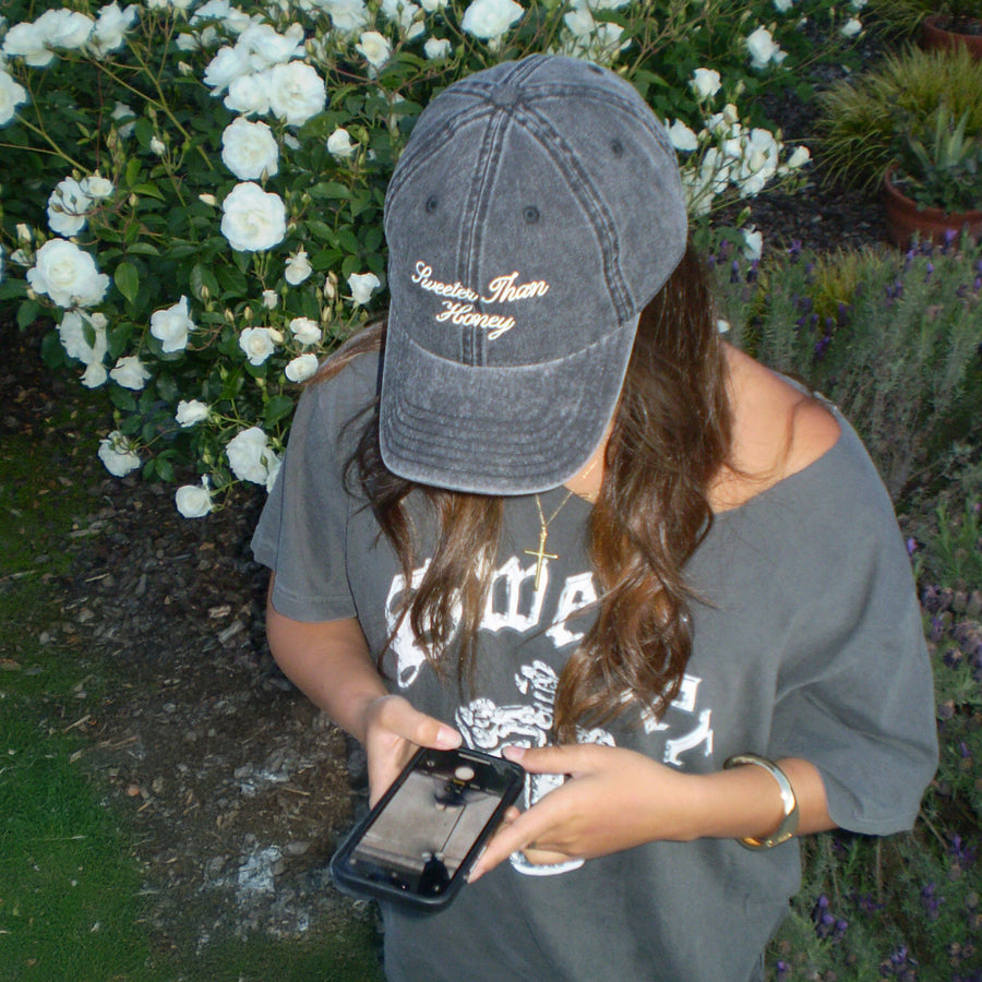 A woman outdoors wearing a dark gray baseball cap with gold embroidered text reading 'Breathe Easy Honey' and a gray graphic T-shirt, holding a smartphone in a garden with white flowers in the background.