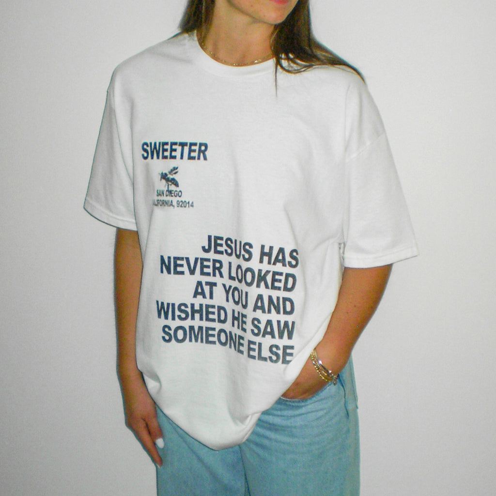 A person wearing a white oversized graphic T-shirt with dark blue text reading 'SWEETER' and a slogan about Jesus, paired with light blue jeans, against a plain light background.