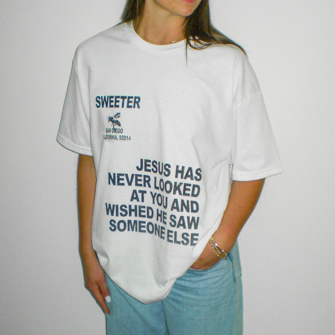 A person wearing a white oversized graphic T-shirt with dark blue text reading 'SWEETER' and a slogan about Jesus, paired with light blue jeans, against a plain light background.