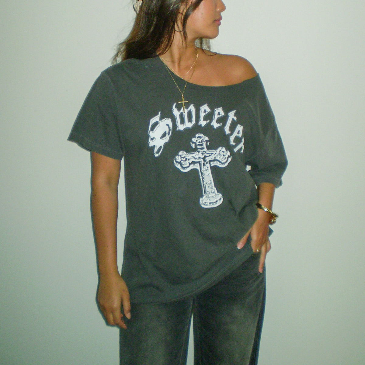 A woman wearing a dark gray off-the-shoulder t-shirt with a white graphic that reads 'Sweeter' above a cross, paired with dark gray jeans, set against a plain light gray wall.