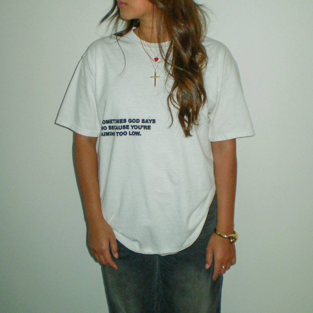 A woman wearing a white crewneck t-shirt with black printed text on the chest, paired with blue jeans, standing against a plain beige wall.
