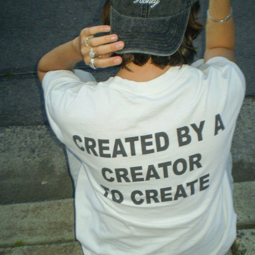 A person viewed from behind wearing a white graphic T-shirt with the text 'CREATED BY A CREATOR TO CREATE' printed in black across the back, along with a dark baseball cap and rings.