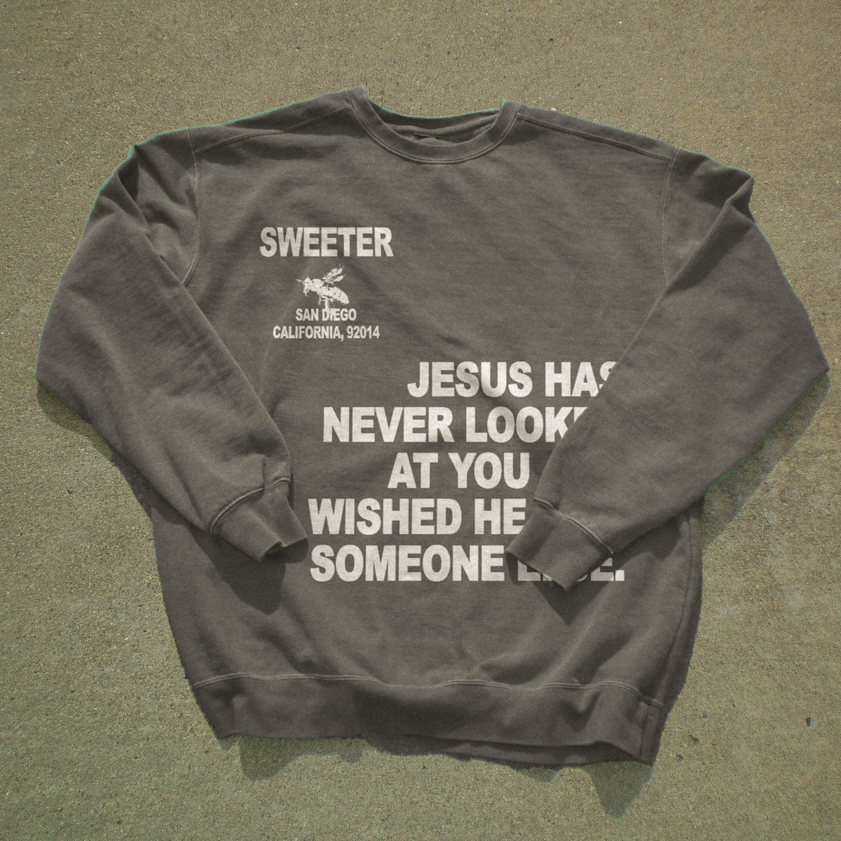 A dark gray crewneck sweatshirt with white printed text and a small graphic, laid flat on a textured gray concrete surface. The text reads 'SWEETER SAN DIEGO CALIFORNIA, 52014' and 'JESUS HAS NEVER LOOKED AT YOU WISHED HE WISHED HE Someone else.