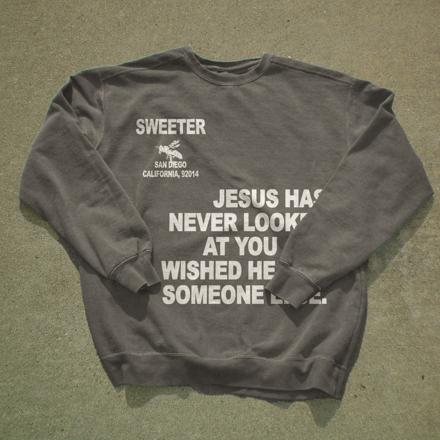 A dark gray crewneck sweatshirt with white printed text and a small graphic, laid flat on a textured gray concrete surface. The text reads 'SWEETER SAN DIEGO CALIFORNIA, 52014' and 'JESUS HAS NEVER LOOKED AT YOU WISHED HE WISHED HE Someone else.