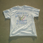 A white short-sleeve graphic t-shirt laid flat on a concrete surface. The shirt features a distressed graphic with the text 'NEVER 2 FLY 2 PRAY' and a hand gesture with a rainbow stripe.