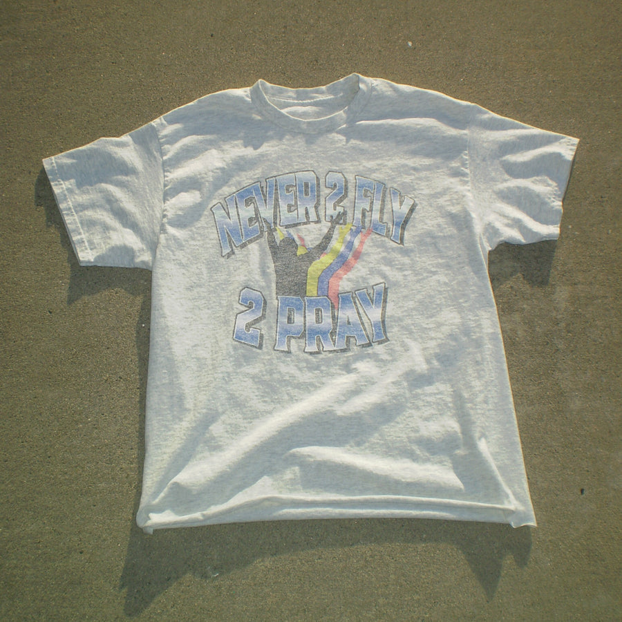 A white short-sleeve graphic t-shirt laid flat on a concrete surface. The shirt features a distressed graphic with the text 'NEVER 2 FLY 2 PRAY' and a hand gesture with a rainbow stripe.