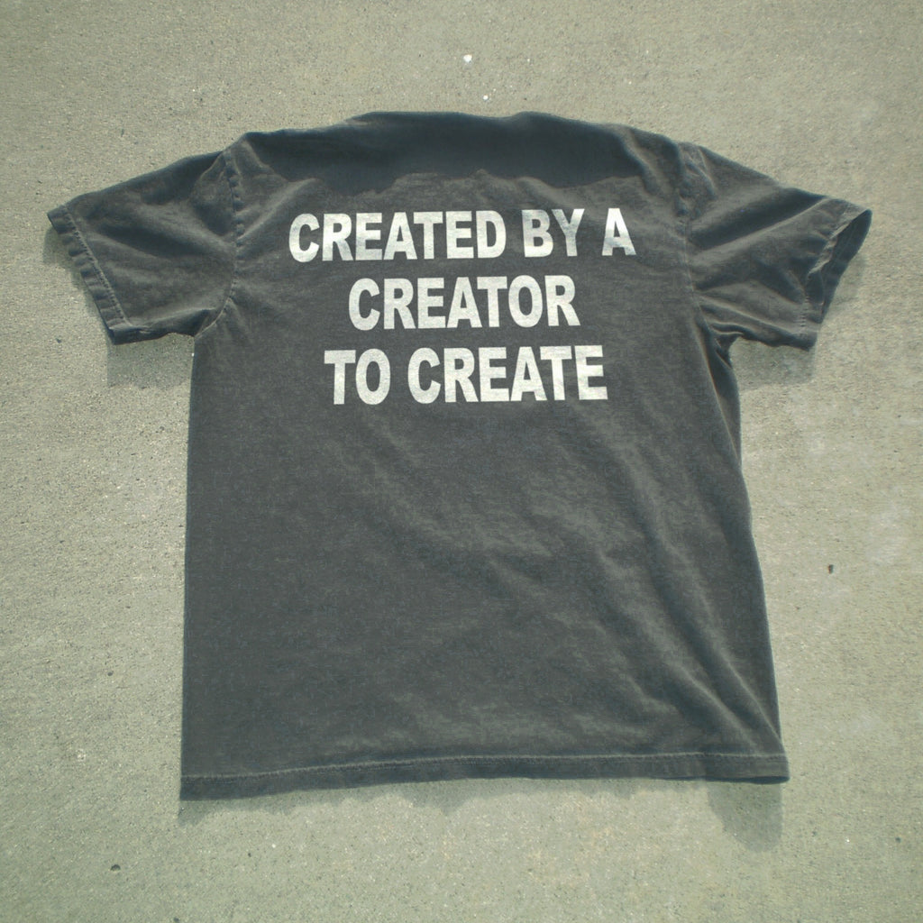 A dark charcoal gray t-shirt laid flat on a textured gray concrete surface, featuring the phrase 'CREATED BY A CREATOR TO CREATE' printed in bold white capital letters across the back.