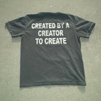 A dark charcoal gray t-shirt laid flat on a textured gray concrete surface, featuring the phrase 'CREATED BY A CREATOR TO CREATE' printed in bold white capital letters across the back.