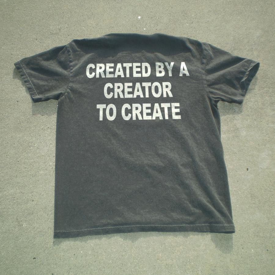 A dark charcoal gray t-shirt laid flat on a textured gray concrete surface, featuring the phrase 'CREATED BY A CREATOR TO CREATE' printed in bold white capital letters across the back.