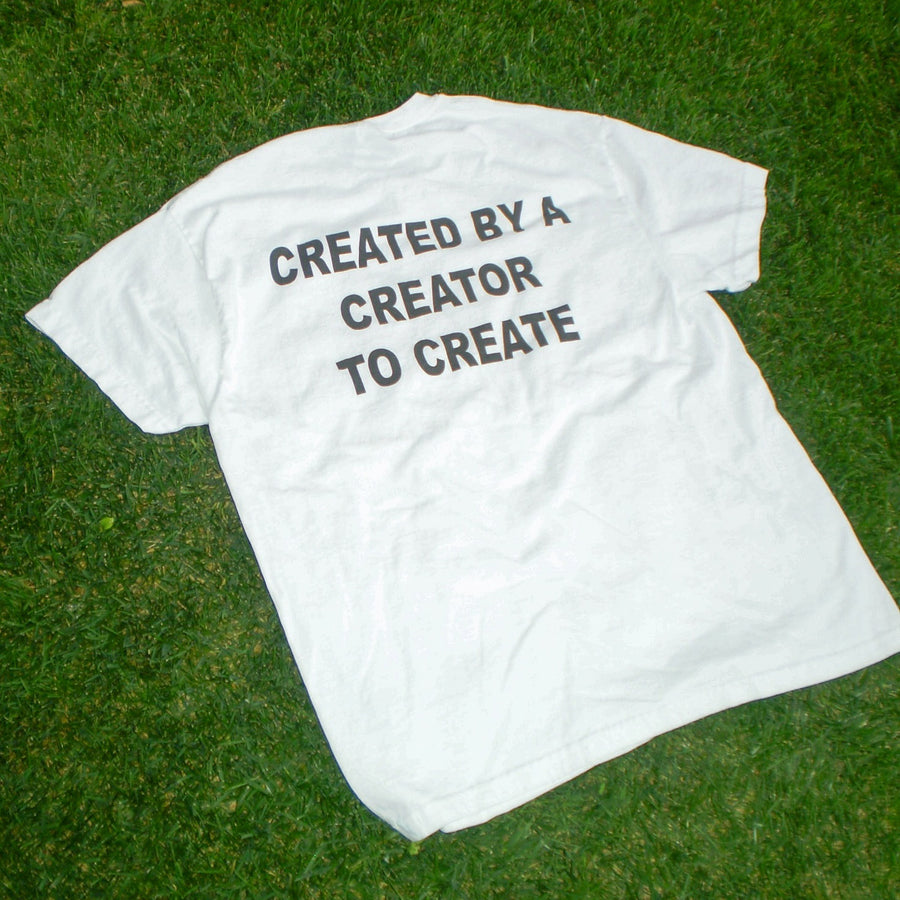 A white short-sleeve t-shirt laid flat on green grass, featuring the text 'CREATED BY A CREATOR TO CREATE' printed in black across the back.
