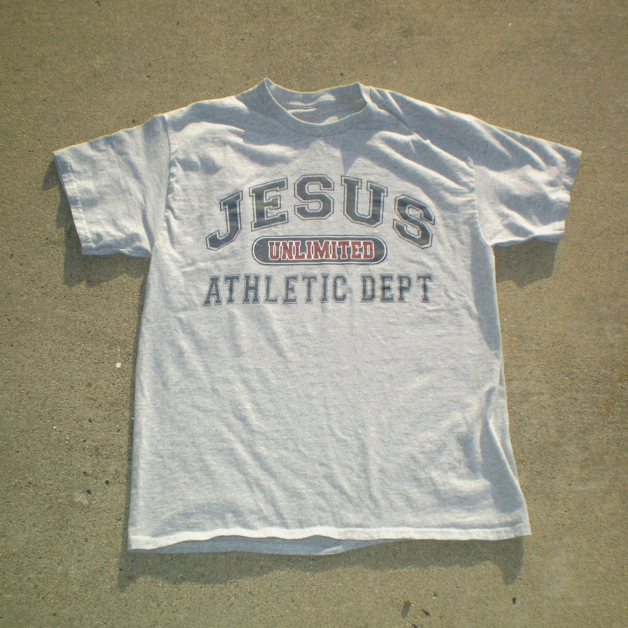 A light gray heather t-shirt laid flat on a sandy surface, featuring a graphic print that reads 'JESUS UNLIMITED ATHLETIC DEPT' in dark gray and red.