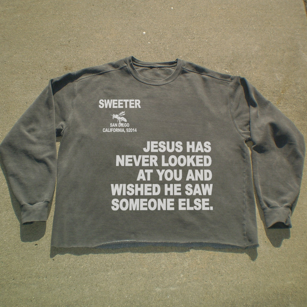 A dark gray long-sleeve t-shirt with white printed text on the front, laid flat on a concrete surface. The text reads 'SWEETER SAN ANGLO CALIFORNIA, 92014' and 'JESUS HAS NEVER LOOKED AT YOU AND WISHED HE SAW SOMEONE ELSE.