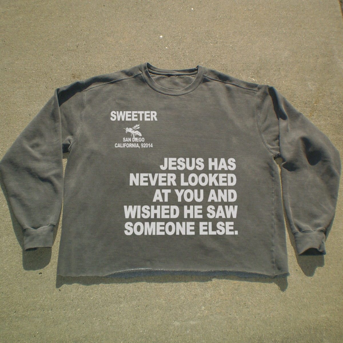 A dark gray long-sleeve t-shirt with white printed text on the front, laid flat on a concrete surface. The text reads 'SWEETER SAN ANGLO CALIFORNIA, 92014' and 'JESUS HAS NEVER LOOKED AT YOU AND WISHED HE SAW SOMEONE ELSE.