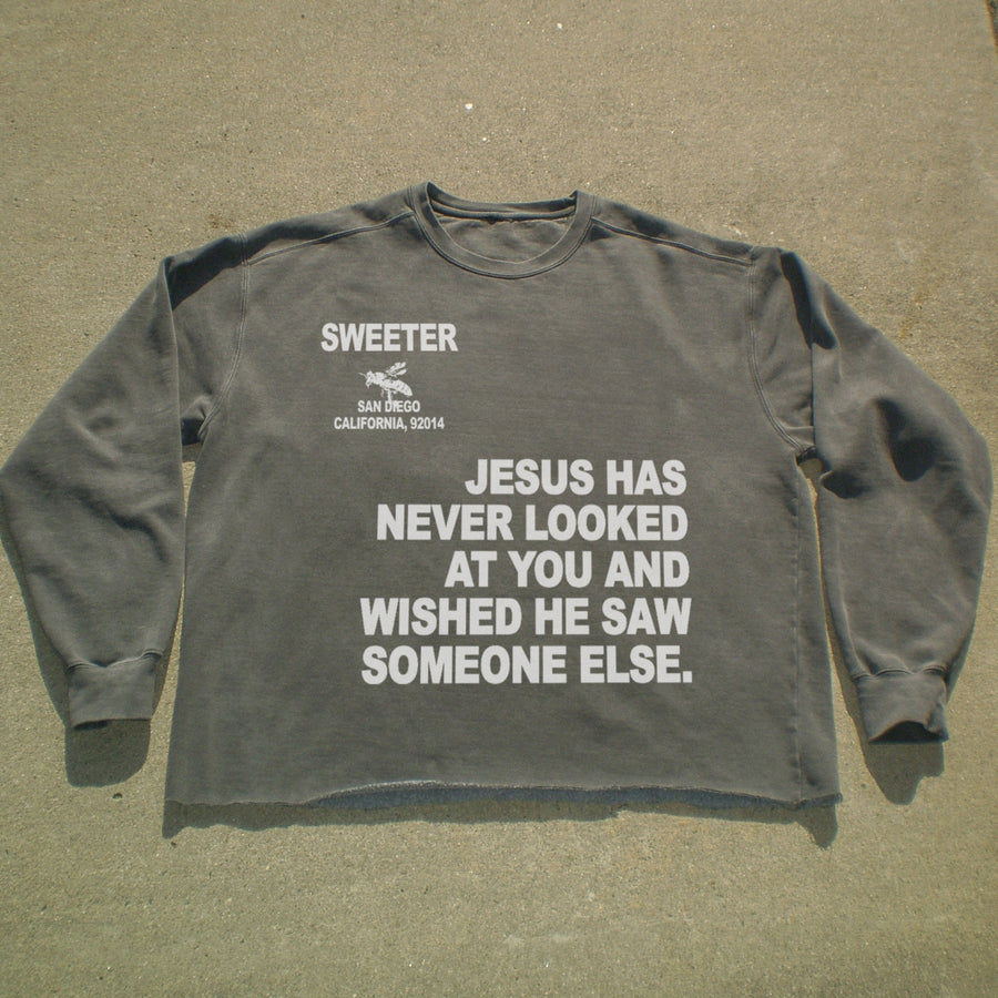 A dark gray long-sleeve t-shirt with white printed text on the front, laid flat on a concrete surface. The text reads 'SWEETER SAN ANGLO CALIFORNIA, 92014' and 'JESUS HAS NEVER LOOKED AT YOU AND WISHED HE SAW SOMEONE ELSE.