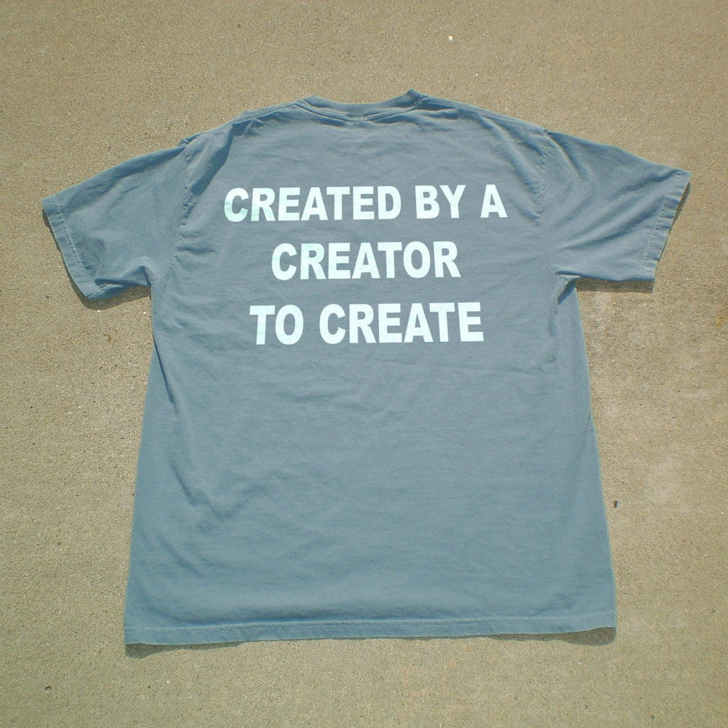 A light blue short-sleeve t-shirt laid flat on a sandy surface, featuring the phrase 'CREATED BY A CREATOR TO CREATE' printed in bold white capital letters across the back.