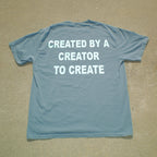 A light blue short-sleeve t-shirt laid flat on a sandy surface, featuring the phrase 'CREATED BY A CREATOR TO CREATE' printed in bold white capital letters across the back.