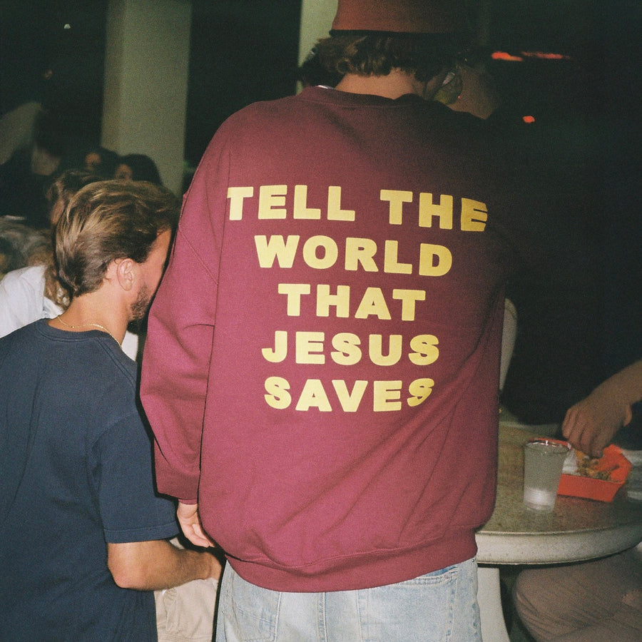 A person standing in a social setting, shown from behind wearing a maroon sweatshirt with the text 'TELL THE WORLD THAT JESUS SAVES' printed in yellow across the back.