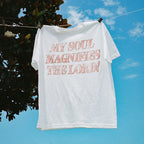 A white short-sleeve t-shirt hanging from a clothesline outdoors, clipped near the shoulders. The shirt has large, distressed pink text on the front that reads, 'MY SOUL MAGNIFIES THE LORD!'.
