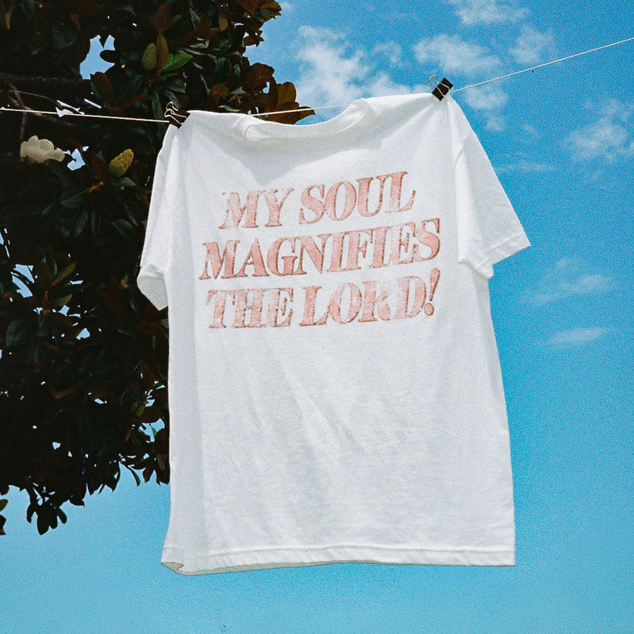 A white short-sleeve t-shirt hanging from a clothesline outdoors, clipped near the shoulders. The shirt has large, distressed pink text on the front that reads, 'MY SOUL MAGNIFIES THE LORD!'.