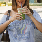 A woman wearing a gray off-the-shoulder graphic t-shirt with the text 'NEVER FLY 2 PRAY' and a rainbow design, holding a green smoothie in a clear cup with a straw.
