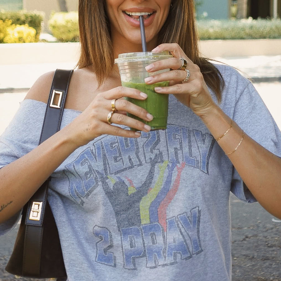 A woman wearing a gray off-the-shoulder graphic t-shirt with the text 'NEVER FLY 2 PRAY' and a rainbow design, holding a green smoothie in a clear cup with a straw.