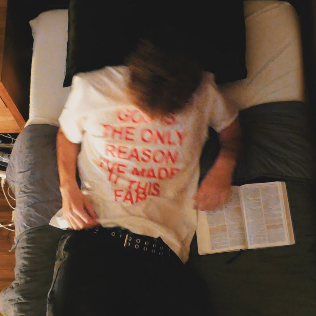 A person lying on a bed wearing a white graphic T-shirt with red text that reads 'GOV'S THE ONLY REASON I'VE MADE IT THIS FAR', with an open book nearby.