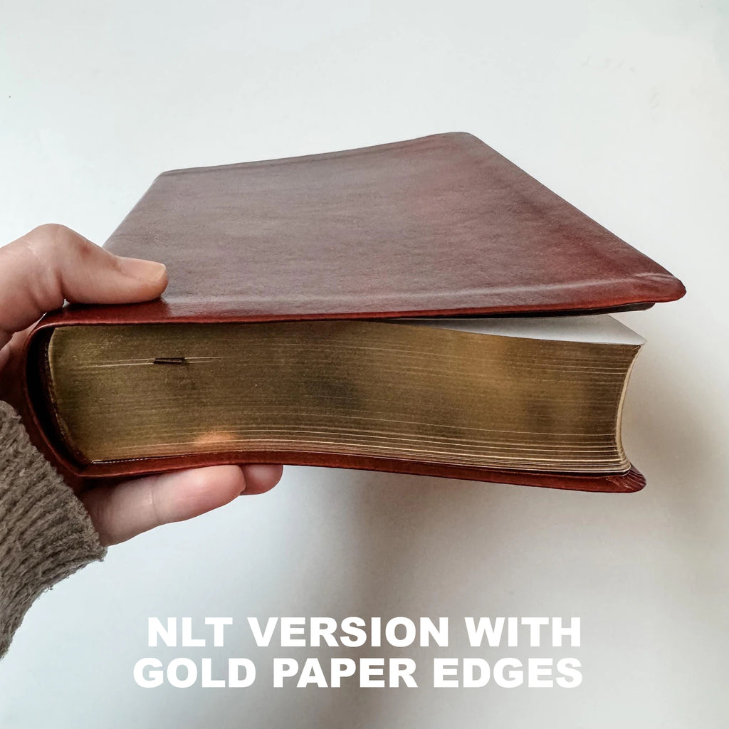 A hand holds a thick book with a reddish-brown leather cover and gold paper edges, with text overlay reading 'NLT VERSION WITH GOLD PAPER EDGES'.