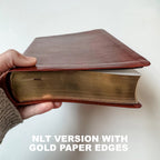 A hand holds a thick book with a reddish-brown leather cover and gold paper edges, with text overlay reading 'NLT VERSION WITH GOLD PAPER EDGES'.