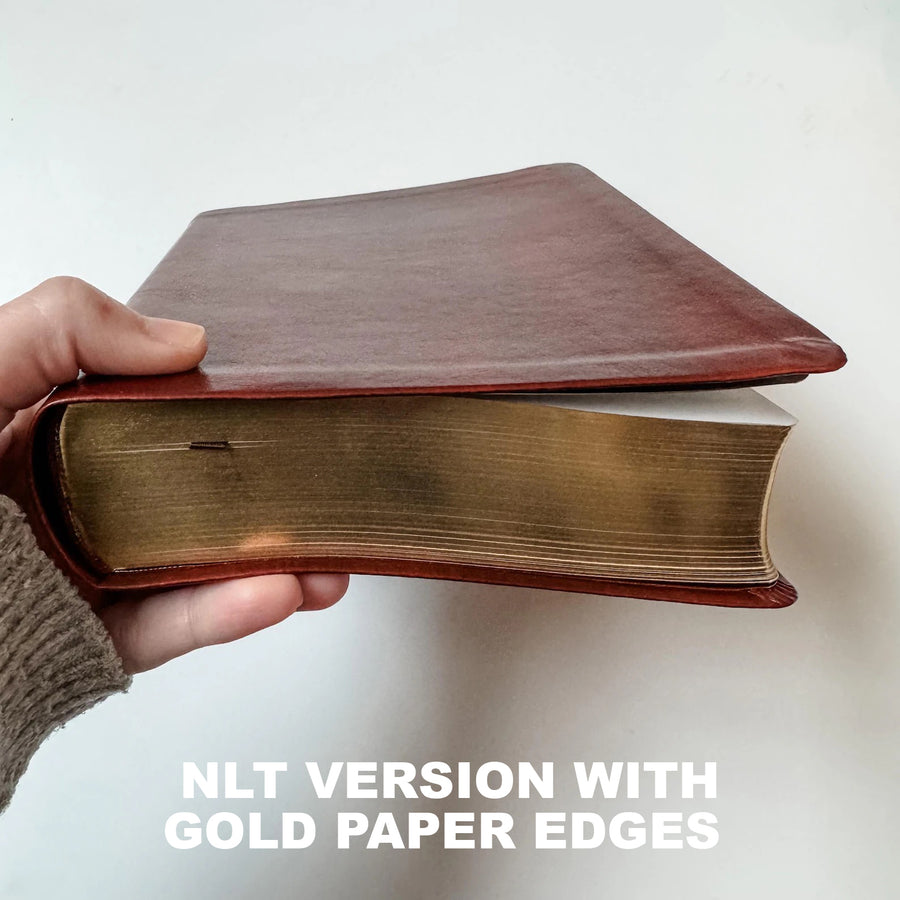 A hand holds a thick book with a reddish-brown leather cover and gold paper edges, with text overlay reading 'NLT VERSION WITH GOLD PAPER EDGES'.
